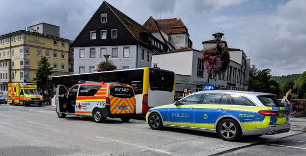 Ein Polizeiwagen neben einem Bus auf einer Straße mit Fußgängern auf dem Gehweg, Gebäuden, Bäumen und einem bewölkten Himmel im Hintergrund.