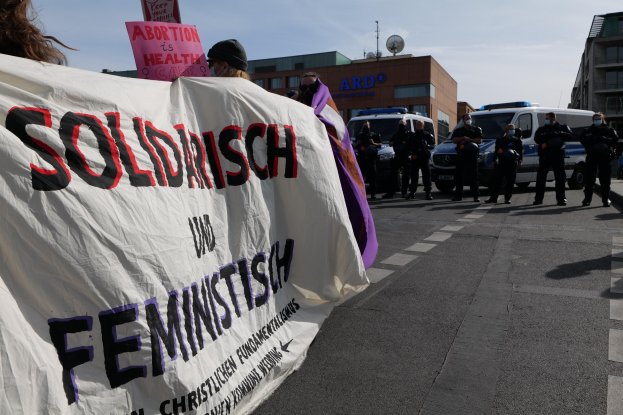 Eine Gruppe von Menschen marschiert auf einer Straße, trägt eine Fahne mit der Aufschrift "Solidarität und Feminismus", mit geparkten Fahrzeugen am Straßenrand und Gebäuden im Hintergrund unter einem klaren blauen Himmel.