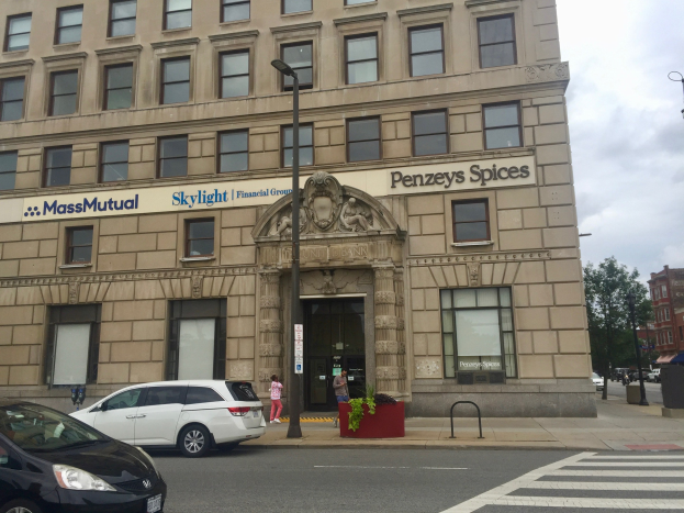 Ein großes Gebäude mit dem Logo der Massachusetts Mutual Financial Group, umgeben von parkenden Autos, Straßenlaternen, beweglichen Fahrzeugen, Fußgängern auf dem Bürgersteig, Topfpflanzen, Bäumen und einem bewölkten Himmel.