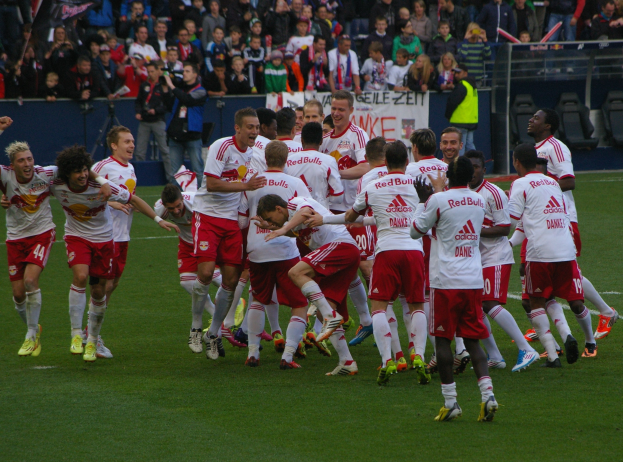 Gruppe von Fußballspielern feiert auf einem Rasenfeld mit Zuschauern in den Rängen, Kameras, Bannern, Geländern und Stühlen im Hintergrund.