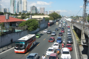 Eine belebte Stadtstraße mit Autos, Bussen und Motorrädern, eine Brücke mit Strommasten und Drähten auf der rechten Seite und Gebäude, Bäume und einen bewölkten Himmel im Hintergrund.