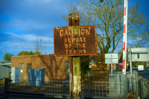 Vorsichtsschild an einem Bahnübergang-Zaun neben Bäumen, Pfählen, einem Gebäude mit Fenstern, Containern, einem Straßenmast, Fahrzeugen auf der Straße und einer bewölkten Himmel.