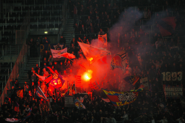 Eine große Menschenmenge in einem Stadion mit Fahnen und Transparenten, beleuchtet von Scheinwerfern, umgeben von Stufen und Geländern, mit einem Feuer in der Mitte.