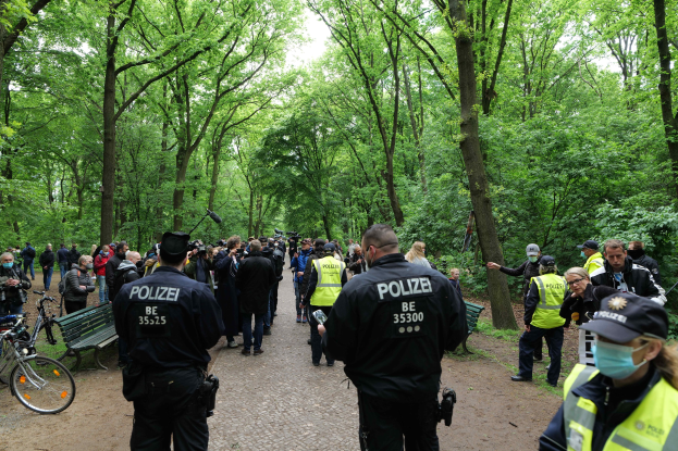 Polizisten vor einer Menge mit Fahrrädern und einer Bank im Vordergrund, Bäume und Himmel im Hintergrund.